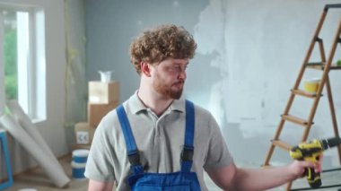 Repairman worker in blue construction overalls is demonstrating how an electric drill works. Portrait of redhead man is posing against backdrop of renovation apartment, ladder, cardboard boxes, window