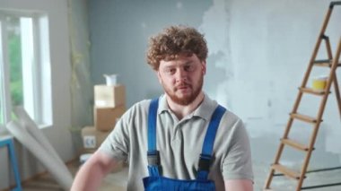 Repairman worker in blue construction overalls is demonstrating how an electric drill works. Portrait of redhead man is posing against backdrop of renovation apartment, ladder, cardboard boxes, window