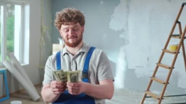 Repairman worker in blue construction overalls joyfully counting money and smiling. Portrait of redhead man posing against backdrop of renovation apartment, ladder, cardboard boxes, window. Concept of