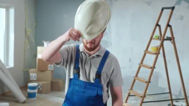 Repairman worker in blue overalls is looking at camera and showing obscene gesture, fuck you. Portrait of redhead man is posing against backdrop of renovation apartment, ladder, cardboard boxes