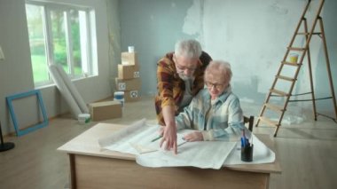 Elderly man and woman are looking through sheet with plan of an apartment and discussing renovation project. Aged couple is sitting at a table and planning the improvement of their home. Concept of