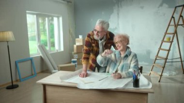 Elderly man and woman are looking through sheet with plan of an apartment and discussing renovation project. Happy aged couple is sitting at a table and planning the improvement of their home. Concept