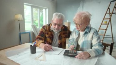 Elderly man and woman are calculating expenses of repairing an apartment on calculator. Aged couple is sitting at a table and planning the improvement of their home. Concept of repair, decoration