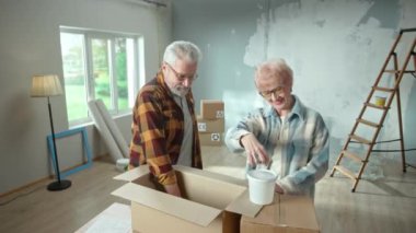 Elderly man and woman are taking out plastic bucket of paint, brush and paint roller from cardboard box. Aged couple is planning repair in apartment and discuss painting the walls. Concept of repair