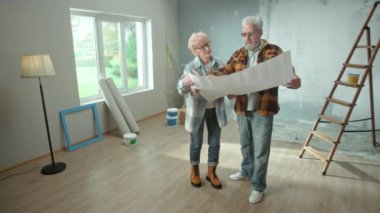 Elderly man and woman are looking through sheet with plan of an apartment and discussing renovation project. Aged couple is planning improvement of their home. Ladder, cardboard boxes and window in