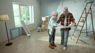 Elderly man and woman are looking through sheet with plan of an apartment and discussing renovation project. Aged couple is planning improvement of their home. Ladder, cardboard boxes and window in
