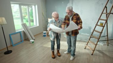 Elderly man and woman are looking through sheet with plan of an apartment and discussing renovation project. Aged couple is planning improvement of their home. Ladder, cardboard boxes and window in
