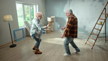 Elderly man and woman are looking through sheet with plan of an apartment and discussing renovation project. Aged couple is having fun and celebrating housewarming. Ladder, cardboard boxes and window