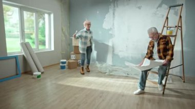 Elderly man and woman are looking through sheet with plan of an apartment and discussing renovation project. Aged couple is having fun and celebrating good start to renovation. Ladder, cardboard boxes