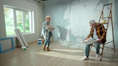 Elderly man and woman are looking through sheet with plan of an apartment and discussing renovation project. Aged couple is having fun and celebrating good start to renovation. Ladder, cardboard boxes