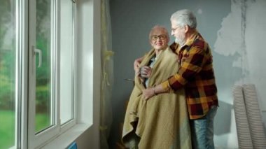 An elderly man wraps plaid around an older woman with cup of tea in his hands. Aged couple of pensioners communicate and share their dreams standing at the window in a cozy home environment