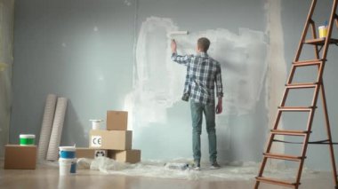 Young man is painting wall with white paint using paint roller. Back view of male painter in checkered shirt enjoying renovations in apartment. Ladder, stack of cardboard boxes, wallpaper and buckets