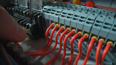 Gray plastic electrical panel with many red wires, yellow bushing ferrules, electrical parts, automatic switches. Close up of hand fasten the wire with a screw using a screwdriver. High voltage