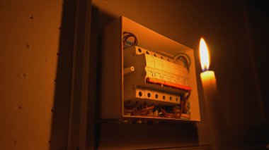 High voltage automatic breaker switch, close up in the dark. Candle illuminates a white electrical panel with many wires, automatic switches, breakers, residual current devices, fuses. Blackout