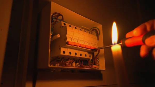 Electrician assembles electrical panel in dark by light of burning candle. Close up of a mans hands tightening a screw with a screwdriver. Blackout. Electric power supply. Switch box with automatic