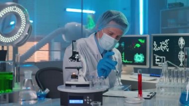 Erkek bir bilim adamı cam pipetle kırmızı sıvı örneği uyguluyor ve mikroskop altında inceliyor. Araştırmacı verileri yazar. Biyokimyasal tulum giyen insanlar...