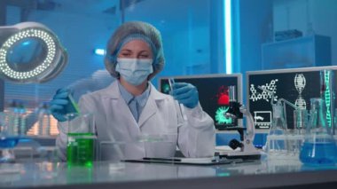 A woman scientist with a pipette draws a green liquid into a test tube, examines it and writes the data of the study. The researcher then examines the sample in Petri dish and writes the study again