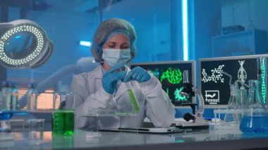 A female scientist using a glass pipette picks up a green liquid and drops it onto a sample in a petri dish. The woman examines it and writes down the data of the study. A researcher works in a modern