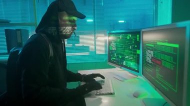 Hacker is typing on a computer keyboard and cracking a password. Access denied, then to access granted. A man in a black hoodie, gloves and a headset is hacking security systems. The spys face