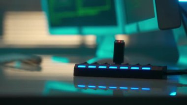 A black flash drive with a red flashing light in the USB port. USB storage in a card reader against the background of computer displays with green lines of codes. A hand in a black glove removes a