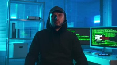Bir hacker 'ın portresi kameraya bakıp başını sallıyor. Siyah kapüşonlu, şapkalı ve kulaklıklı bir adam bilgisayarlı bir ofisin önünde poz veriyor. Monitörlerde yeşil çizgiler var.