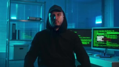 Bir hacker 'ın portresi kameraya özgüvenle bakıyor ve kollarını kavuşturuyor. Siyah kapüşonlu ve şapkalı bir adam bilgisayarlı bir ofisin önünde poz veriyor. Monitörlerde program kodunun yeşil çizgileri var.