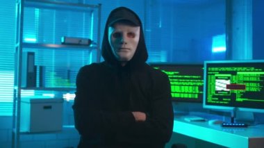 Portrait of a hacker in a white mask confidently looking at the camera and crosses his arms. A man in a black hoodie and cap poses in front of an office with computers. On the monitors are green lines