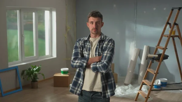 Portrait of man crossing his arms and looking seriously at camera. Young confident male in checkered shirt and posing against backdrop of room in process of being renovated. Concept of repair