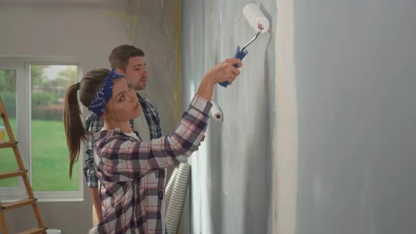 Young couple is painting wall with white paint using paint rollers on window backdrop. Pretty woman and man in checkered shirts enjoying renovations in apartment. Concept of repair, finishing work