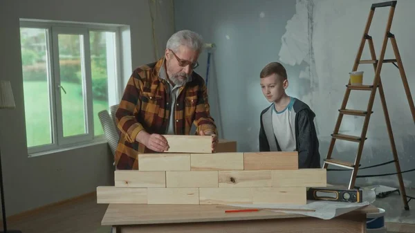 Grandpa and grandson are building a wall of wooden blocks on the table. An elderly man teaches a teenager the basics of construction and repair. The concept of family, joint renovation in apartment