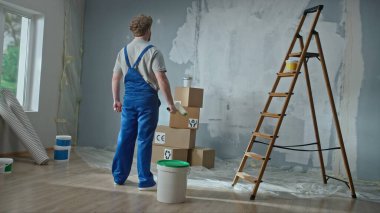 Rear view of a foreman or house painter in blue construction overalls. A red haired man with a paint roller in his hands plans to paint the walls in the apartment. The concept of repair, finishing