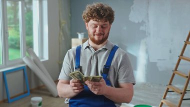 Repairman worker in blue overalls is counting money and smiling. Portrait of a redhead man is posing against backdrop of apartment, ladder, cardboard boxes, window. Concept of repair, finishing works