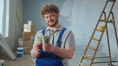 Repairman worker in blue overalls is counting money and smiling. Portrait of a redhead man is posing against backdrop of apartment, ladder, cardboard boxes, window. Concept of repair, finishing works