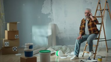 Tired elderly man is sitting on a ladder and calling a master to repair an apartment using a mobile phone. Aged pensioner solved problem with help of the master. Concept of repair, decoration