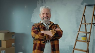Elderly gray haired man with beard is looking at camera, crossing arms over his chest and smiling. Portrait of pensioner is posing in an apartment against background of wall painted with white paint