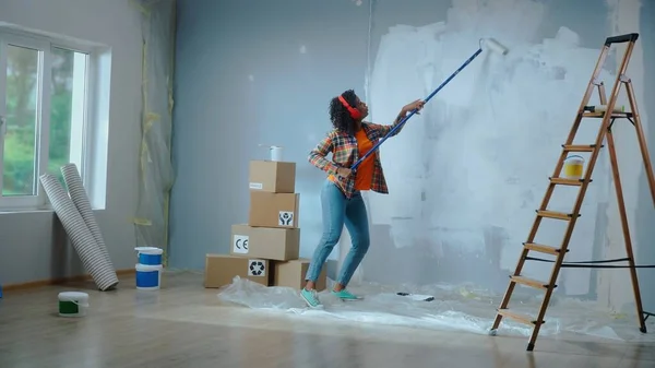 African American woman in red headphones paints wall with white paint using long paint roller and enjoy the music. Black female making repairs in an apartment. Window, ladder, cardboard boxes. Concept
