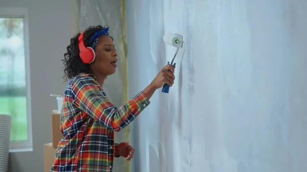 African American woman in red headphones paints the wall with white paint using paint roller and enjoy the music. Black woman is making repairs in an apartment. Concept of repair, finishing works