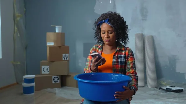 Upset African American woman holding blue bowl into which drops of water are dripping from ceiling leak. Female calling on a mobile phone to master repairman or plumber and asking for help in repairs