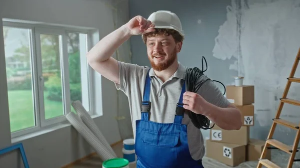 Foreman or electrician in blue overalls, white helmet and coil of black cable in hands. Redhead man is planning repairs in an apartment against backdrop of ladder, cardboard boxes and window. Concept