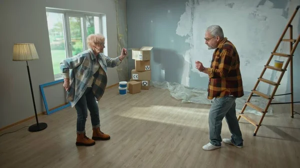 Elderly man and woman dance cheerfully and smile. A couple of pensioners have fun and celebrate housewarming. Ladder, cardboard boxes and a framed window. The concept of repair, decoration, interior