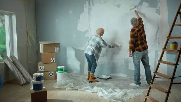 Elderly man and woman are painting wall with white paint using paint rollers. Couple of pensioners are making repairs to their apartment, in the background of window, stepladder, cardboard boxes