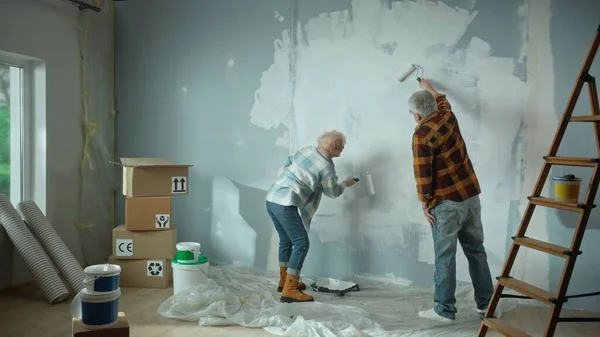 Elderly man and woman are painting wall with white paint using paint rollers. Couple of pensioners are making repairs to their apartment, in the background of window, stepladder, cardboard boxes