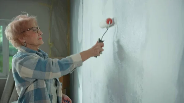 Elderly woman is painting wall with white paint using paint roller. Female pensioner is making repairs to their apartment against the backdrop of a window with bright sunlight. Side view. Concept of
