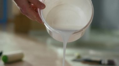 The hands of an unrecognizable person pour white paint from a bucket into a black container. A jet of paint pours into a container, spills and fills it. Close up. Preparation for the repair and