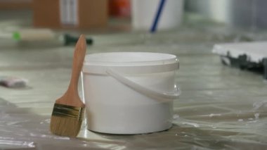 A round plastic bucket with a handle and brush stand on the floor covered with oilcloth. Close up of a white pail with paint and a clean wooden brush against a blurred background. The concept of