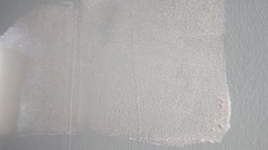 The unrecognizable person paints a gray wall with white paint using a paint roller. A close up of a round, blue handled roller slides over a wall and paints over it. The concept of repair, painting