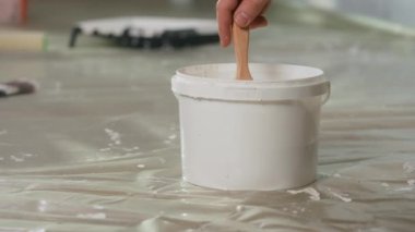The hand of an unrecognizable man dips a brush into a bucket of white paint and stirs it. Then he takes out a brush in paint and puts it on the floor covered with oilcloth. Close up. The concept of
