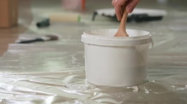 The hand of an unrecognizable man takes a brush and dips it into a plastic bucket of white paint. Close up of a bucket of paint on an oilcloth covered floor. The concept of repair, painting, finishing