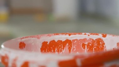 The hand of an unrecognizable man dips a brush into a orange bucket of white paint and stirs it. A man wipes paint from a yellow brush on the edge of the rim. Close up. The concept of repair, painting