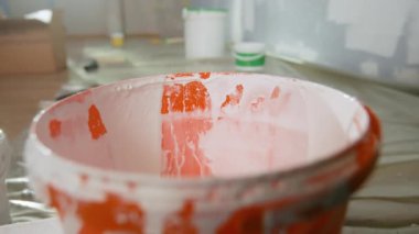 The hand of an unrecognizable man dips a brush into a orange bucket of white paint and stirs it. A man wipes paint from a yellow brush on the edge of the rim. Close up. The concept of repair, painting